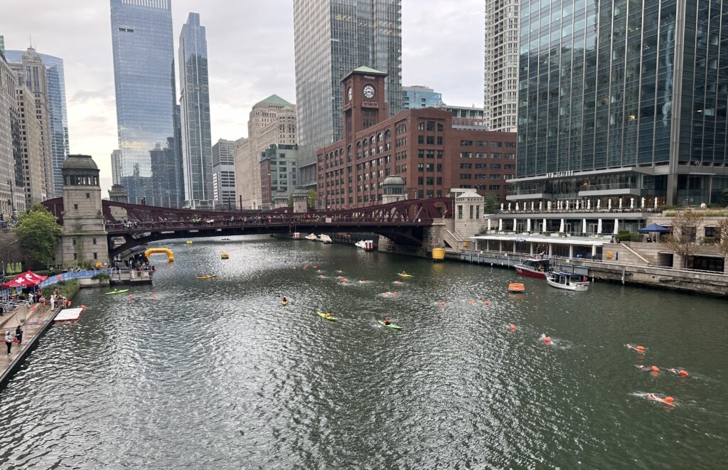 Uma vista ampla mostra uma linha de nadadores no rio Chicago se aproximando de uma ponte, edifícios que se elevam sobre a beira da água