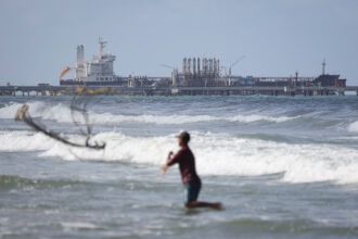 Um pescador lança uma rede ao mar enquanto um petroleiro é visto ancorado ao fundo em 18 de dezembro de 2025, em Puerto Cabello, Venezuela. Crédito: Jesus Vargas/Getty Images