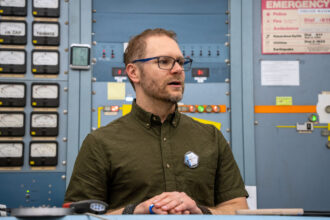 Nicholas Spada está em frente a um painel de instrumentos no Laboratório Nuclear Crocker da UC Davis com um monitor de exposição à radiação preso no bolso de sua camisa em 25 de março.