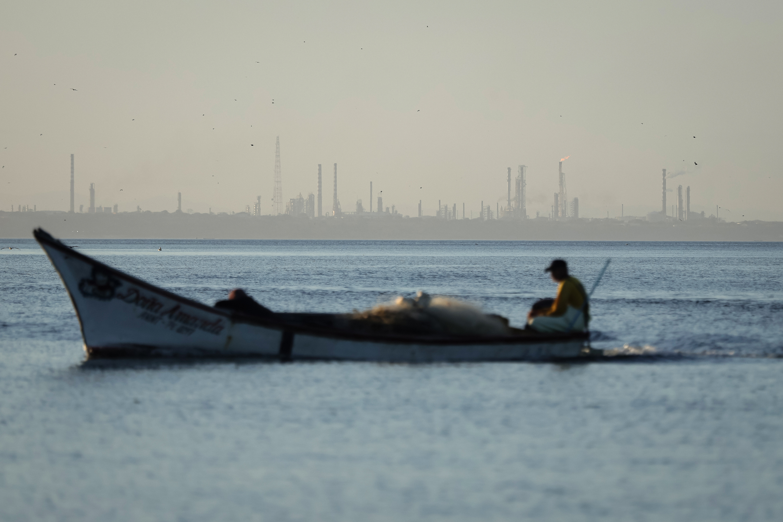 The Punta Cardon oil refinery is seen in the background of a fisherman sailing his boat from a beach in Amuay, Venezuela. Credit: Jesus Vargas/picture alliance via Getty Images