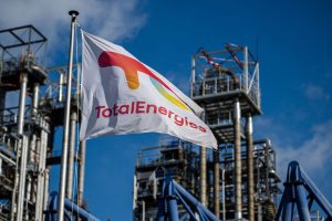 A TotalEnergies flag flies in the foreground, with a refinery in the background.