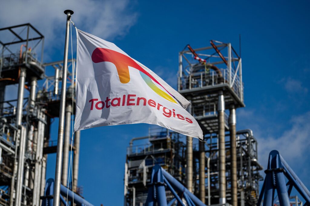 A TotalEnergies flag flies in the foreground, with a refinery in the background.