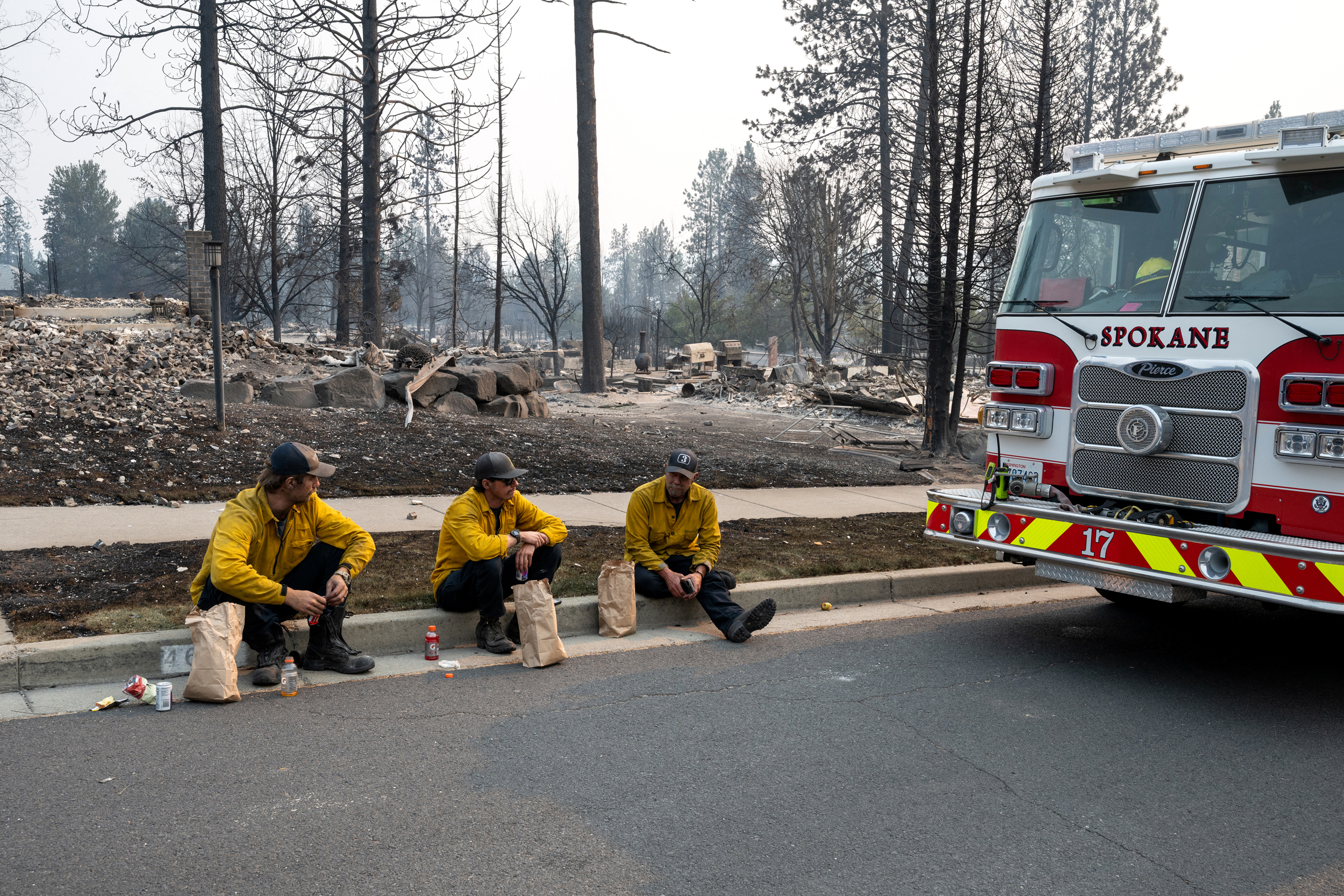 Membros do Corpo de Bombeiros de Spokane sentam-se no meio-fio em frente a casas residenciais queimadas no bairro de Balboa, em Spokane, em 2 de agosto. Crédito: Erick Doxey/AFP via Getty Images